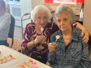 An old lady with her arm around another old lady. Both are holding lollipops.